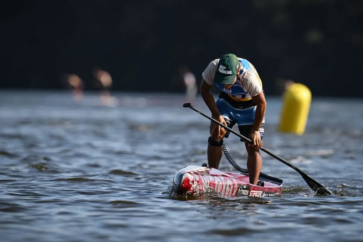 A male racer in a green cap leans over his paddle on a red race board near a yellow buoy