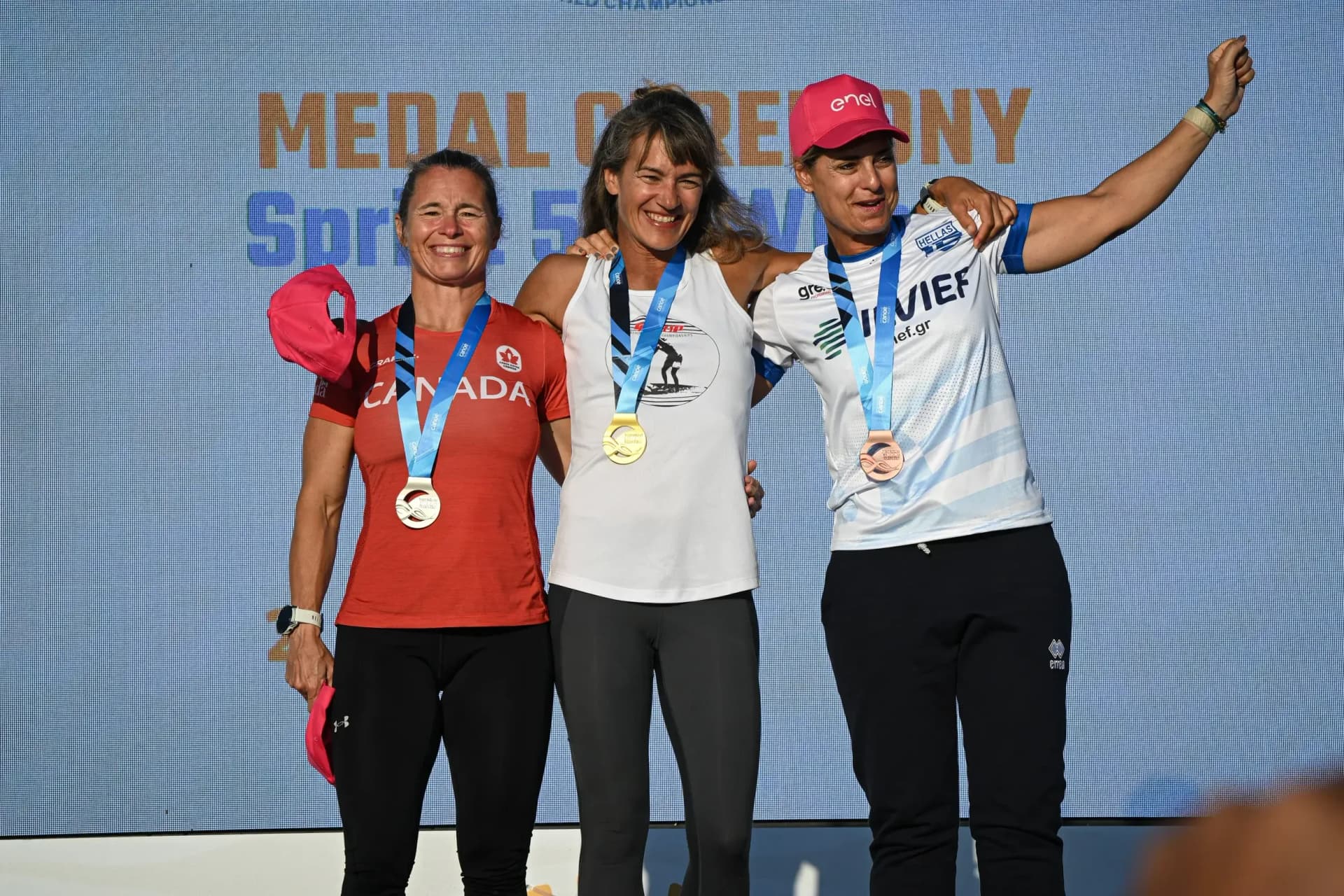 Three women stand together on the medal podium in front of a medal ceremony backdrop, the gold medalist in the middle