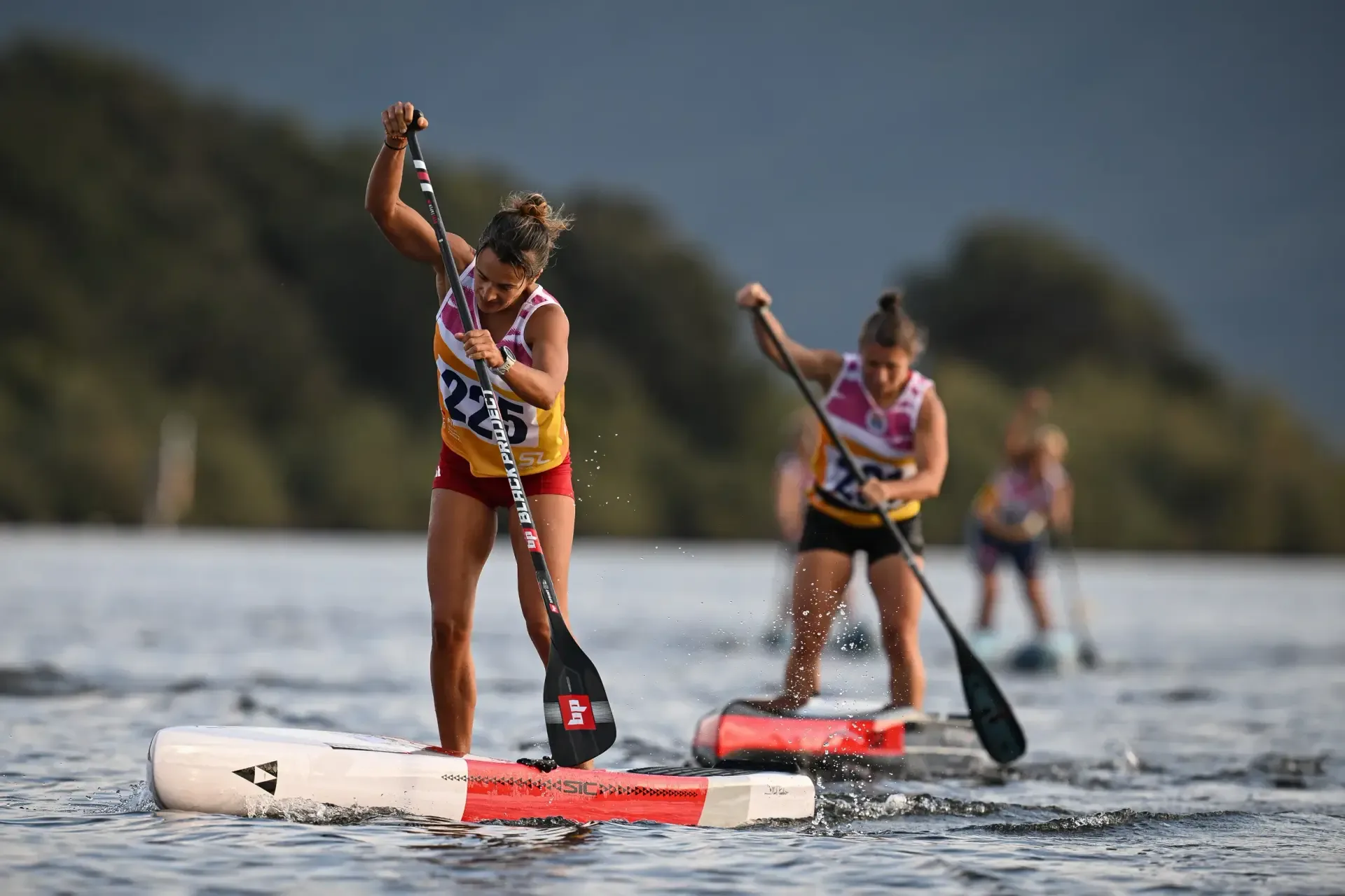 A racer in bib 225 digs in with her paddle on a red and white race board, another racer close behind