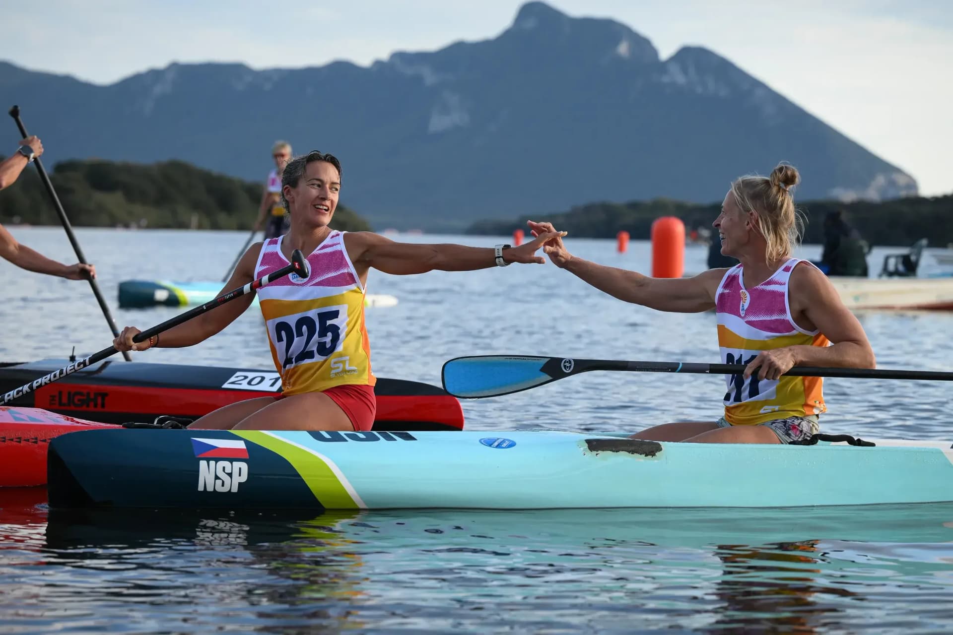 Two racers kneeling on their boards reach across to touch hands after a race, a mountain rising behind the water