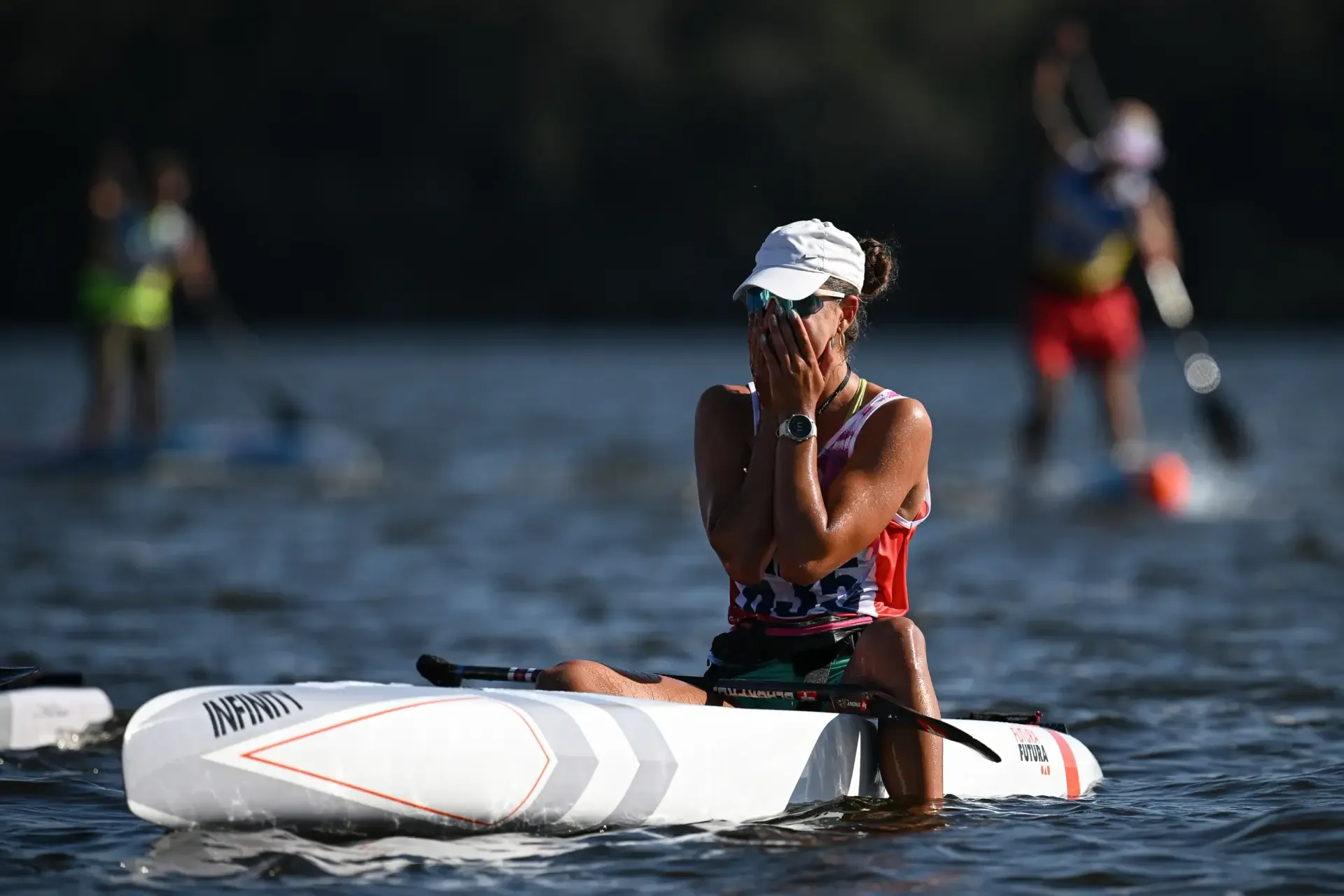 A racer in a white cap sits on her white race board and covers her face with both hands after the finish, other paddlers blurred behind her