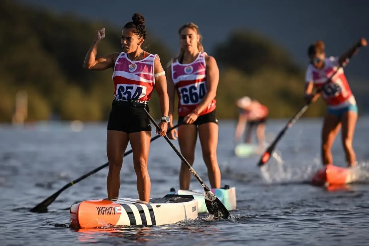A racer with a Puerto Rico flag on her race board raises a fist on the water, two other racers paddling behind her