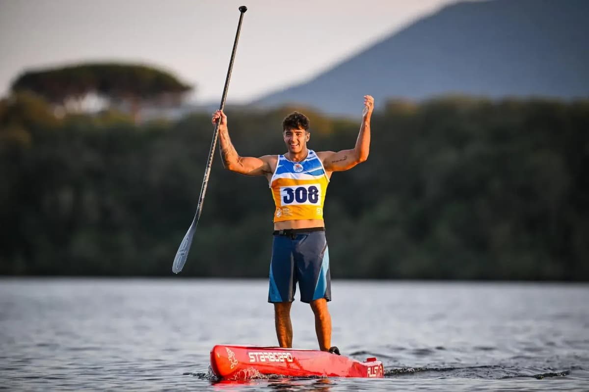 A male racer in bib 308 stands on a red race board with both arms raised and his paddle held high, trees and a mountain behind him