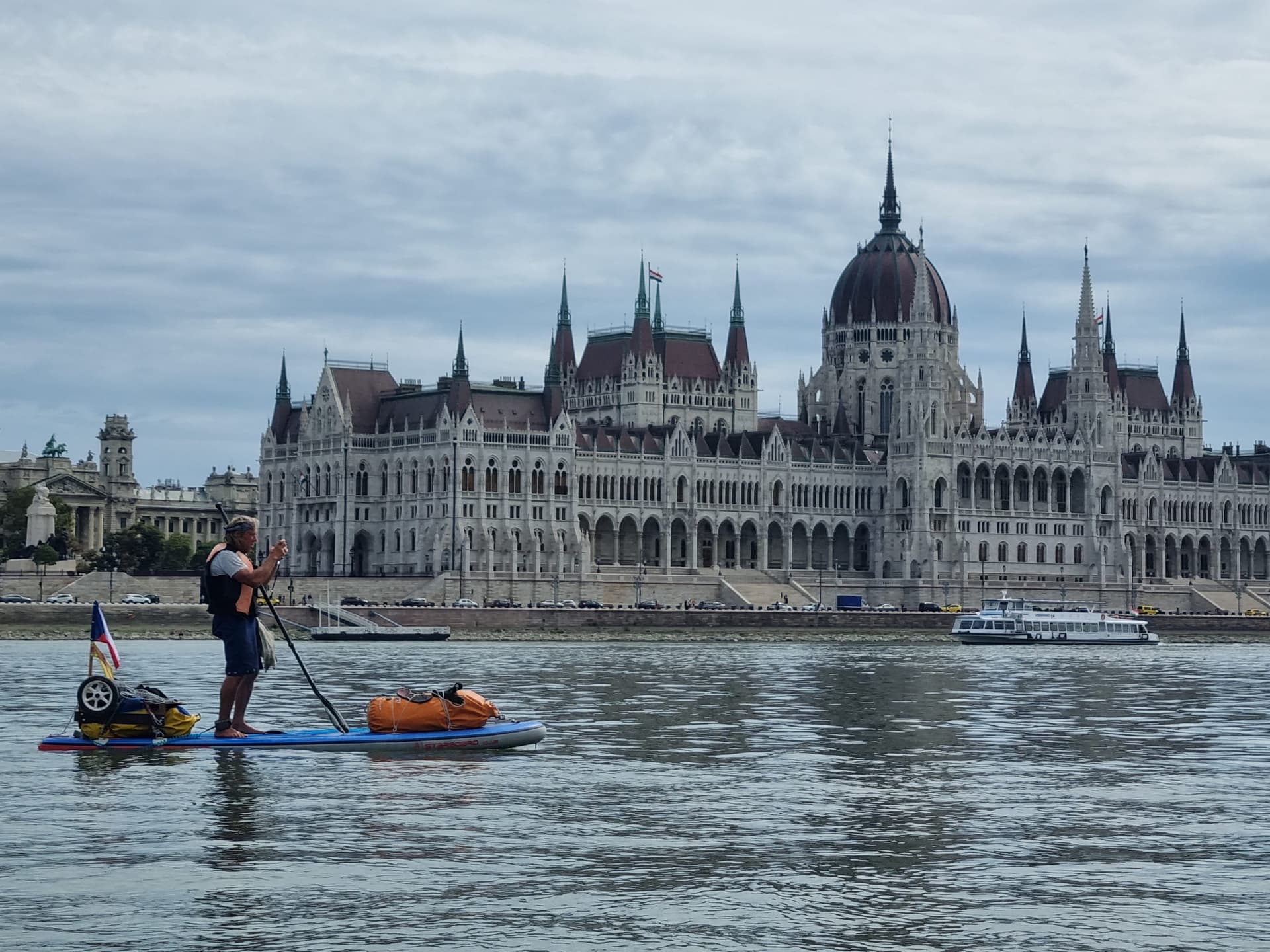 A paddler with a loaded touring board, flag and cart wheels stands on a wide river in front of an ornate domed parliament building