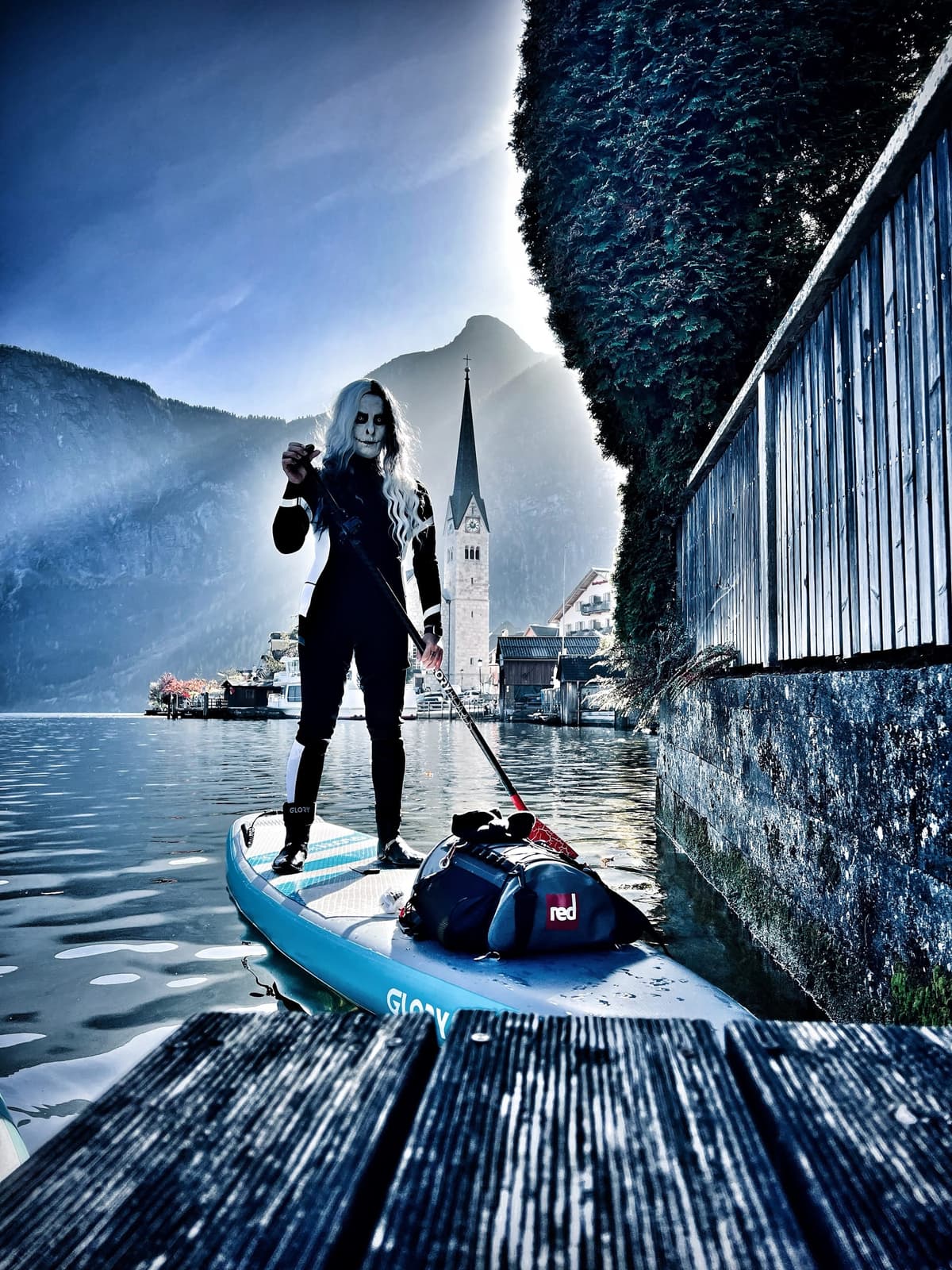 A paddler in skull face paint and a wetsuit stands on a board beside a jetty, with a church spire and mountains behind in hazy light
