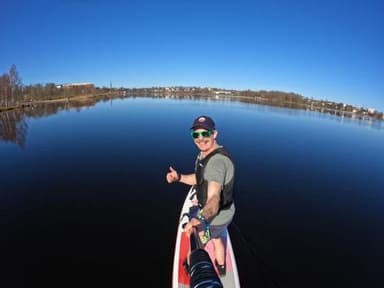 A smiling paddler in sunglasses and a life vest gives a thumbs up on a glassy lake under a clear blue sky