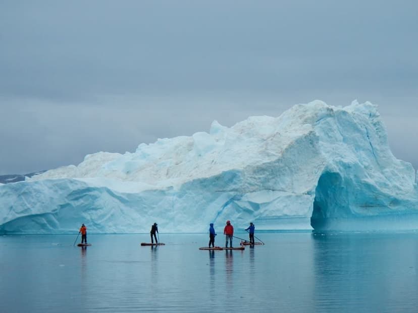 Five paddlers stand on boards on calm water in front of a huge iceberg with an arch