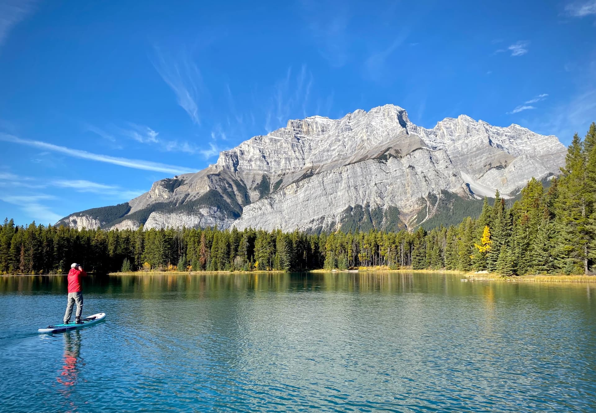 A paddler in a red jacket stands on a board on a calm lake below a rocky mountain and pine forest